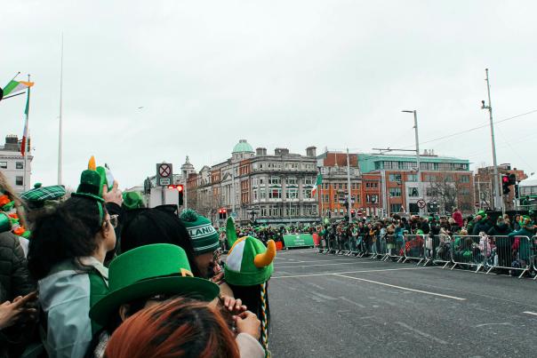 A large crowd of St. Patrick's Day parade spectators lining a city street, many wearing green leprechaun hats, shamrock accessories, and green costumes, with Irish flags waving above the crowd behind metal barricades on an overcast day.