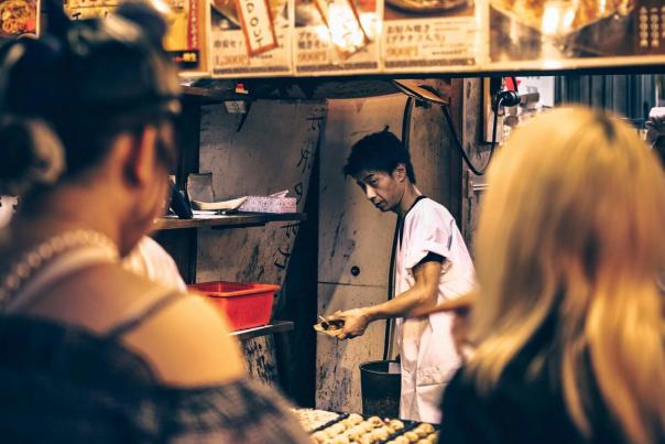 A vendor preparing food at a busy night market street stall with colorful dishes on display