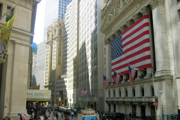 Looking down Wall Street at Nassau Street with the New York Stock Exchange's neoclassical columns and American flags visible on the right, pedestrians filling the street below