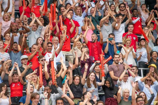 Large crowd of soccer fans cheering and celebrating inside a packed stadium during an international football match with red team jerseys and rally sticks visible throughout the stands.