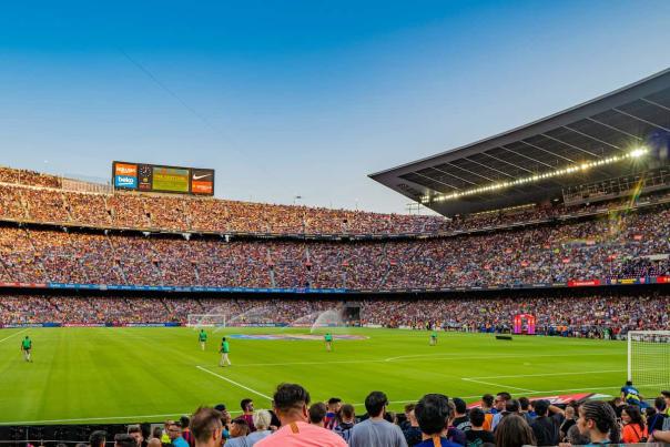 A wide-angle view of a packed soccer stadium at sunset, with a full crowd, bright green pitch, and grounds crew on the field.