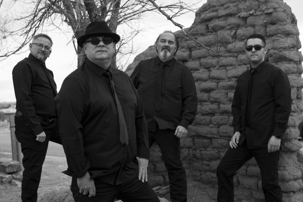 Four members of Sangre Joven stand in front of a crumbling adobe wall in black outfits.