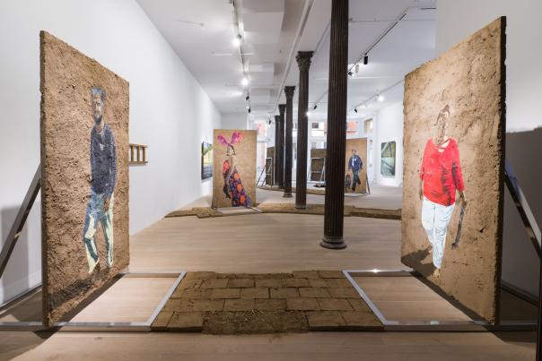 A contemporary gallery installation features large textured panels resembling earthen walls, each painted with full‑body figures, arranged along a central path of brick-like tiles under bright overhead lighting.