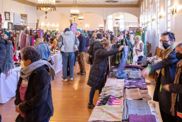 A bustling indoor craft fair, with people in winter clothing browsing colorful textile displays.