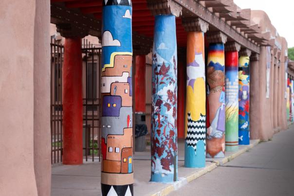 A row of adobe-style columns painted with colorful murals, including geometric patterns, landscapes, and abstract designs, lines a covered walkway beside a stucco building.