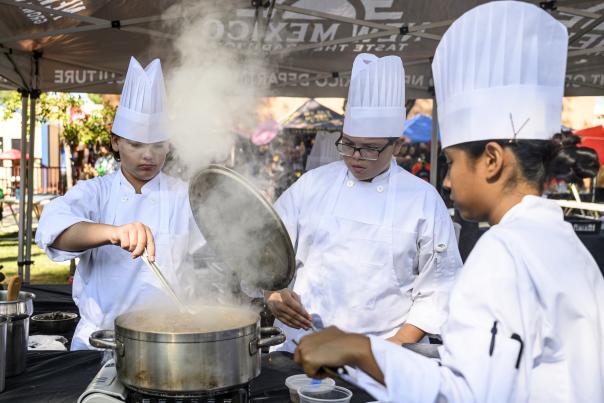 High school culinary students in chef hats prepare green chile stew during the New Mexico State Fair cooking competition.