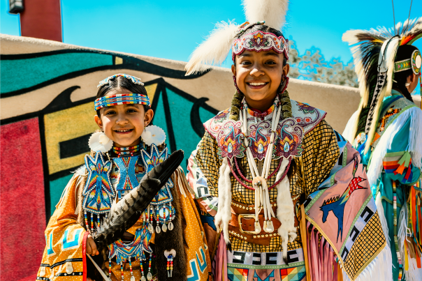 Two smiling dancers in colorful traditional regalia stand side by side at the New Mexico State Fair's Indian Village.