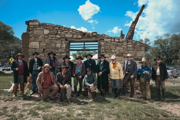 Group of people dressed in Old West–style clothing posing in front of a partially collapsed stone building under a bright blue sky.