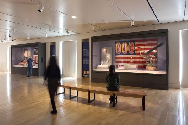 People explore a museum gallery with large display cases featuring historic artifacts and an American flag backdrop.