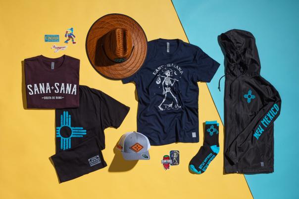 A flat lay of New Mexico–themed apparel and accessories displays graphic T‑shirts, a wide‑brimmed hat, a jacket, socks, a cap, and colorful stickers arranged on a split yellow and turquoise background.