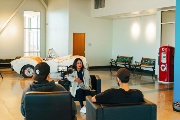 A camera crew films an interview in a spacious room that features a vintage white convertible car, several benches, and a red Texaco gas pump.