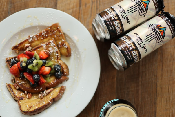 A plate of french toast topped with fruit and powdered sugar sits next to two cans of French Toast Stout and the top of a glass of stout.
