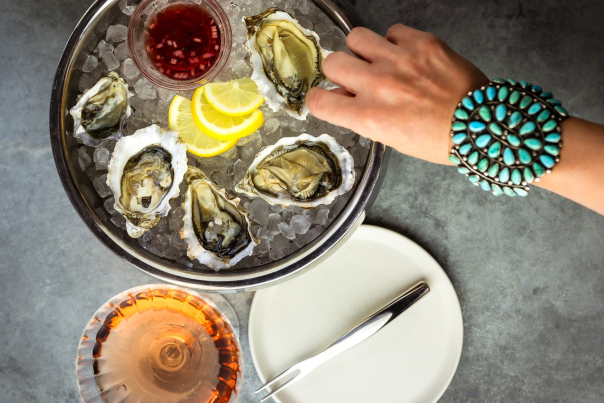 A hand wearing a turquoise bracelet reaches for a plate of six oysters on ice with lemon slices. A small dish of red sauce, glass of rosé, and fork are nearby.