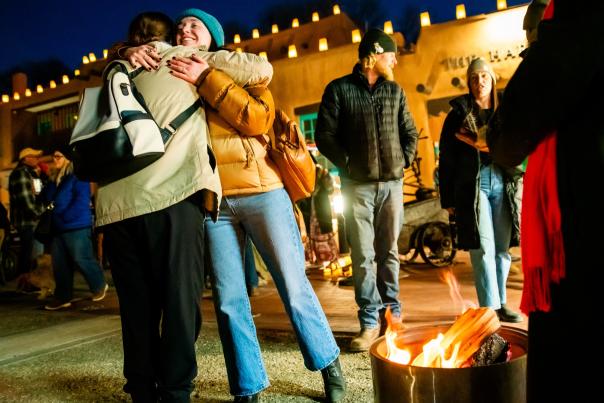 Two people warmly embrace in a festive outdoor setting with a fire pit and soft lights.