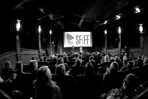 A black and white photo of an audience in a theater facing a screen that says "SFIFF" as two people stand on stage.