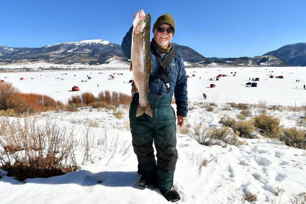 A man in winter clothing showcases a large fish caught during ice fishing. Snowy landscape with red tents and distant mountains under a clear blue sky.