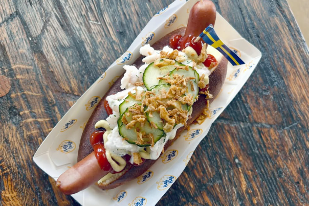 A hot dog topped with ketchup, mustard, cucumbers, chopped onions, and a Swedish flag toothpick in a tray on a rustic wooden table.