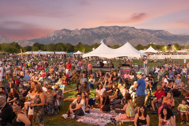 A large outdoor crowd sits on blankets and folding chairs at a festival on a grassy field, with white event tents scattered throughout and a mountain range glowing in the pink sunset sky behind them.