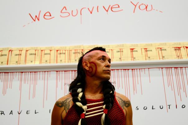 Indigenous man with braided hair and face paint stands solemnly.
