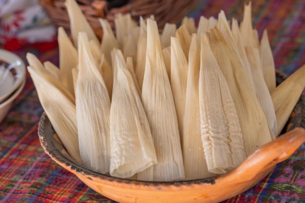 A terracotta bowl filled with dried corn husks sits on a colorful tablecloth.