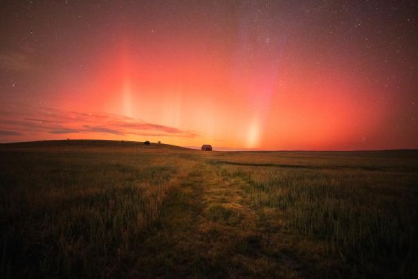 A barn sits on the horizon of a grassy field beneath a vibrant red aurora borealis.