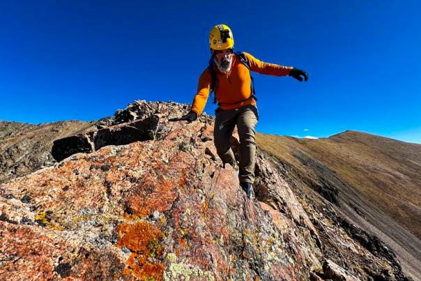 A climber in an orange shirt and yellow helmet ascends a rocky ridge, balancing carefully. The sky is clear blue, and the terrain is rugged and uneven.