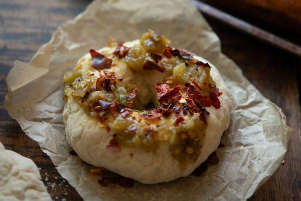A bagel topped with chopped green chiles and red pepper flakes rests on brown parchment paper.