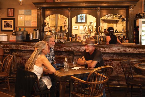 Three people sit at a wooden table in a dimly-lit, old-timey saloon.