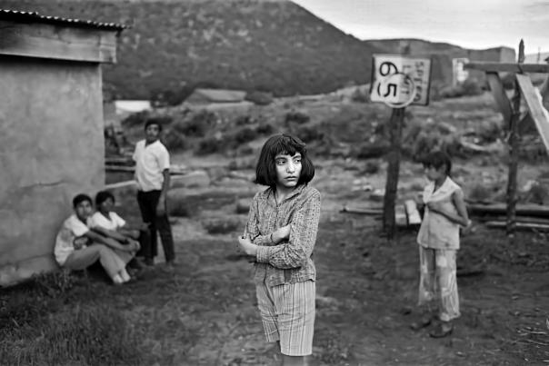 Black and white photograph of a young girl standing with folded arms in the foreground beside a dirt road in Petaca, New Mexico.