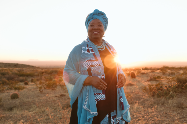 Woman in a blue patterned dress and headscarf stands in a dry field at sunset.