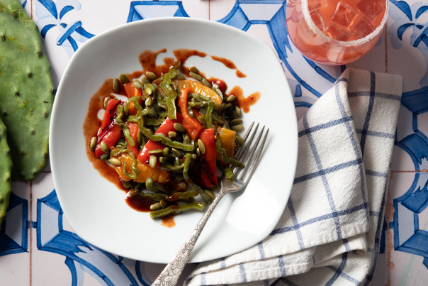 Plate of nopales salad with vegetables and a pink prickly pear margarita beside a cactus pad.