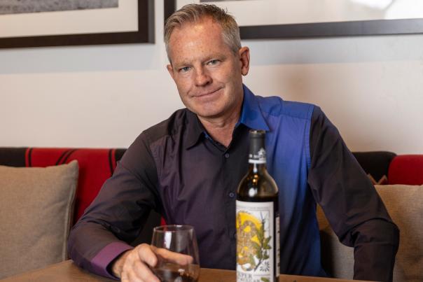 A man in a black and blue button-down shirt sits at a table in front of a bottle of wine and a glass of wine.
