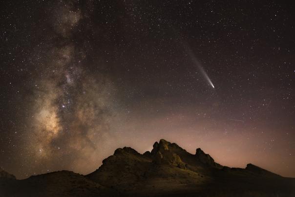 Starry night sky over a silhouetted mountain range, featuring a bright comet with a long tail and the Milky Way galaxy visible.