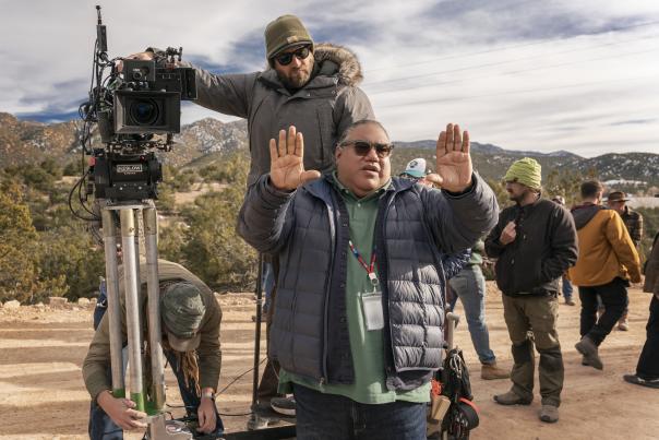 A film crew member in a blue puffer jacket holds up both hands in a 'stop' gesture while standing in front of a camera on a dirt road.