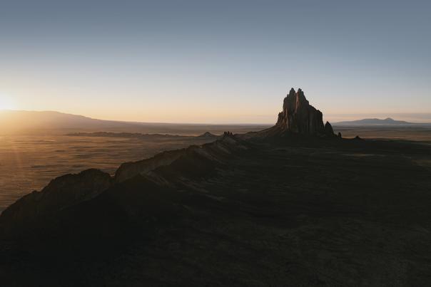 A dramatic volcanic rock formation rises sharply from a vast desert plain at sunrise, casting long shadows across the landscape beneath a clear, pale sky.
