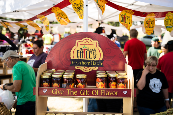 Farmers’ market booth displaying jars of chile products from The Fresh Chile Co., with shoppers in the background.