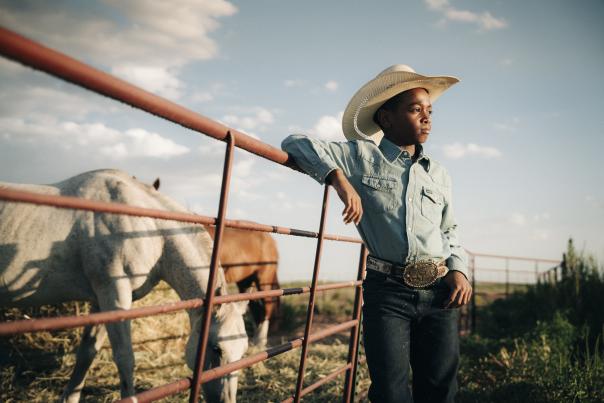 Young bull rider Parker Hooks in cowboy hat and denim shirt leaning on a ranch fence in Hobbs, New Mexico.