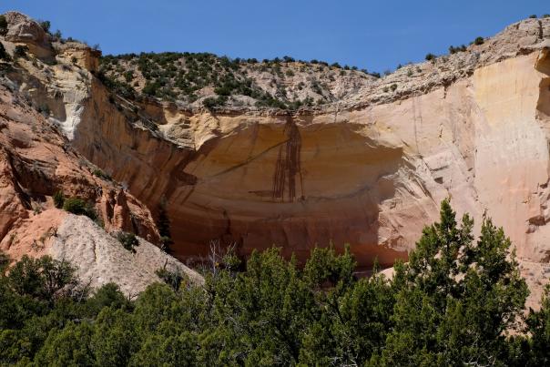 A dramatic sandstone cliff with layered red, orange, and cream rock rises above a line of green shrubs under a clear blue sky.