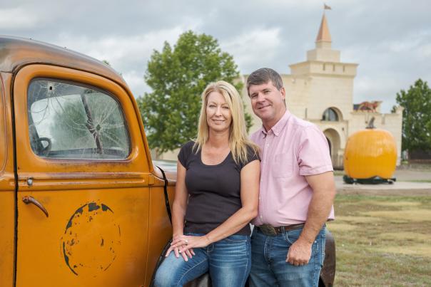 A man and a woman stand in front of an orange vintage truck, with a large, sculpted pumpkin and a castle-like building in the background.