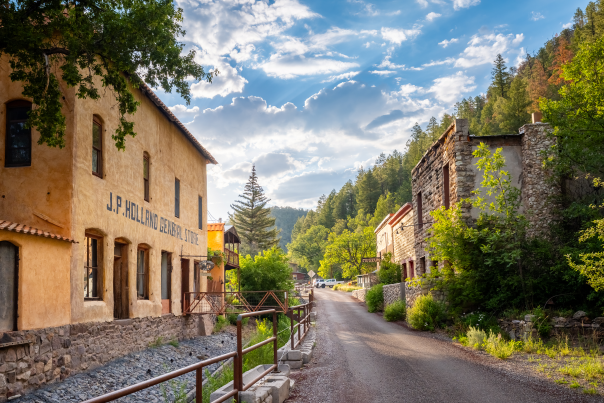 Historic buildings line the narrow main street of Mogollón, New Mexico, under a bright blue sky surrounded by forested canyon walls.