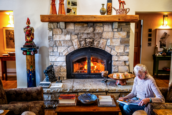 Cozy living room with a warm stone fireplace, books, and decorative items. A person sits reading on a chair.