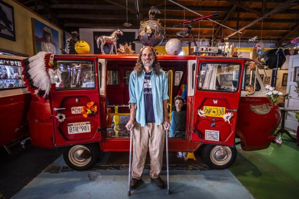 Person standing with crutches in front of a brightly decorated red van inside an eclectic, art-filled space.