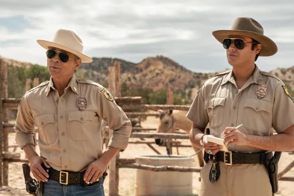 Two men in sheriff uniforms and sunglasses stand outside in a sunlit desert setting, with a horse in the background.