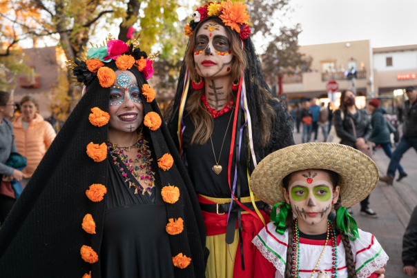 Three people, two women and a girl, pose in traditional Dia de los Muertos attire and makeup.