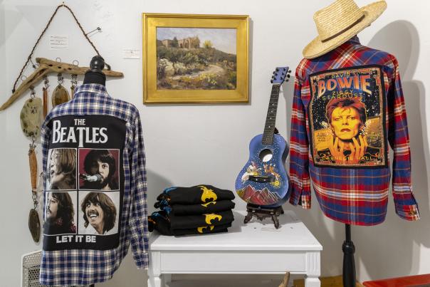 Blue acoustic guitar decorated with bright floral designs next to a red flannel shirt embroidered with a David Bowie design on the back.