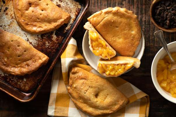 Flaky, golden-brown empanaditas filled with peach filling are arranged on a baking sheet and a plate.
