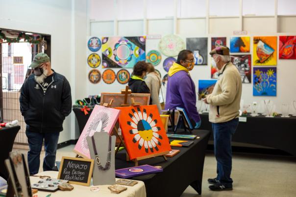 People wearing masks browse colorful artwork displayed on the walls and tables of an indoor art market.