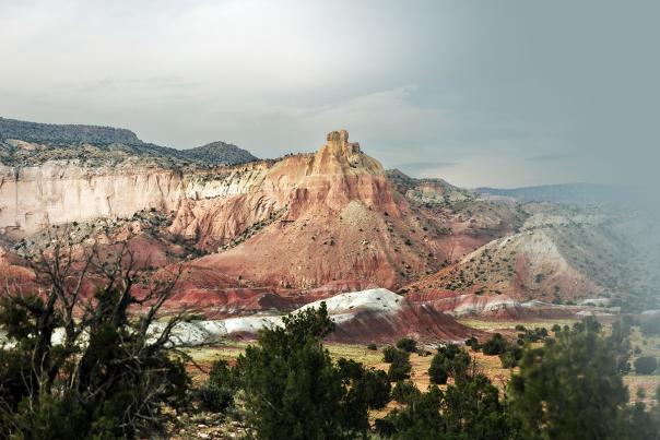 Multicolored desert cliffs and buttes rise above rolling red, white, and tan hills dotted with shrubs under an overcast sky.