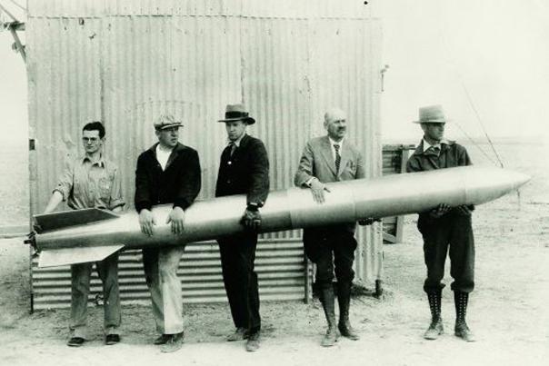 Five men hold a large, cylindrical rocket horizontally in front of a corrugated metal building.