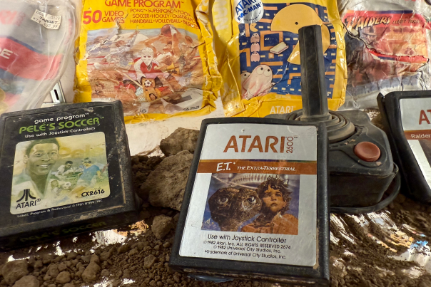 A collection of vintage Atari 2600 game cartridges lies on the ground, partially buried in dirt. Visible titles include "Pele's Soccer" and "E.T. the Extra-Terrestrial." A nearby joystick is also visible.